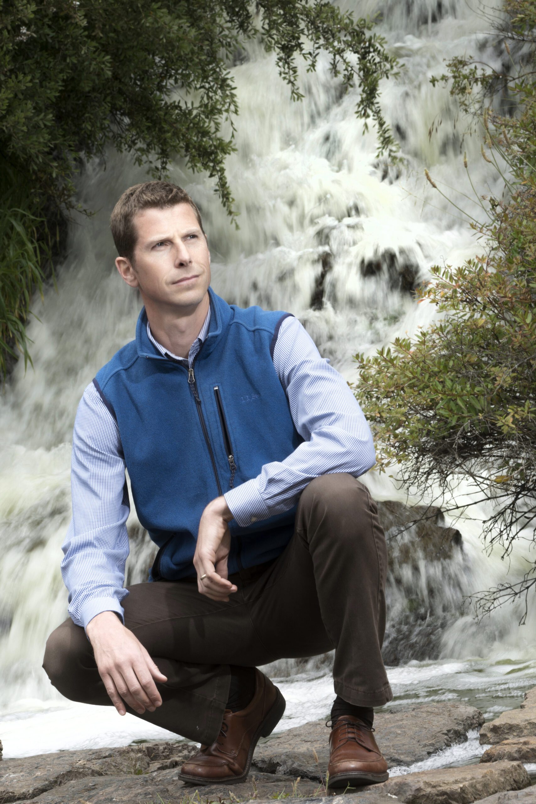 Man wearing a blue vest, light blue dress shirt, brown slacks and brown shoes, crouching in front of a waterfall