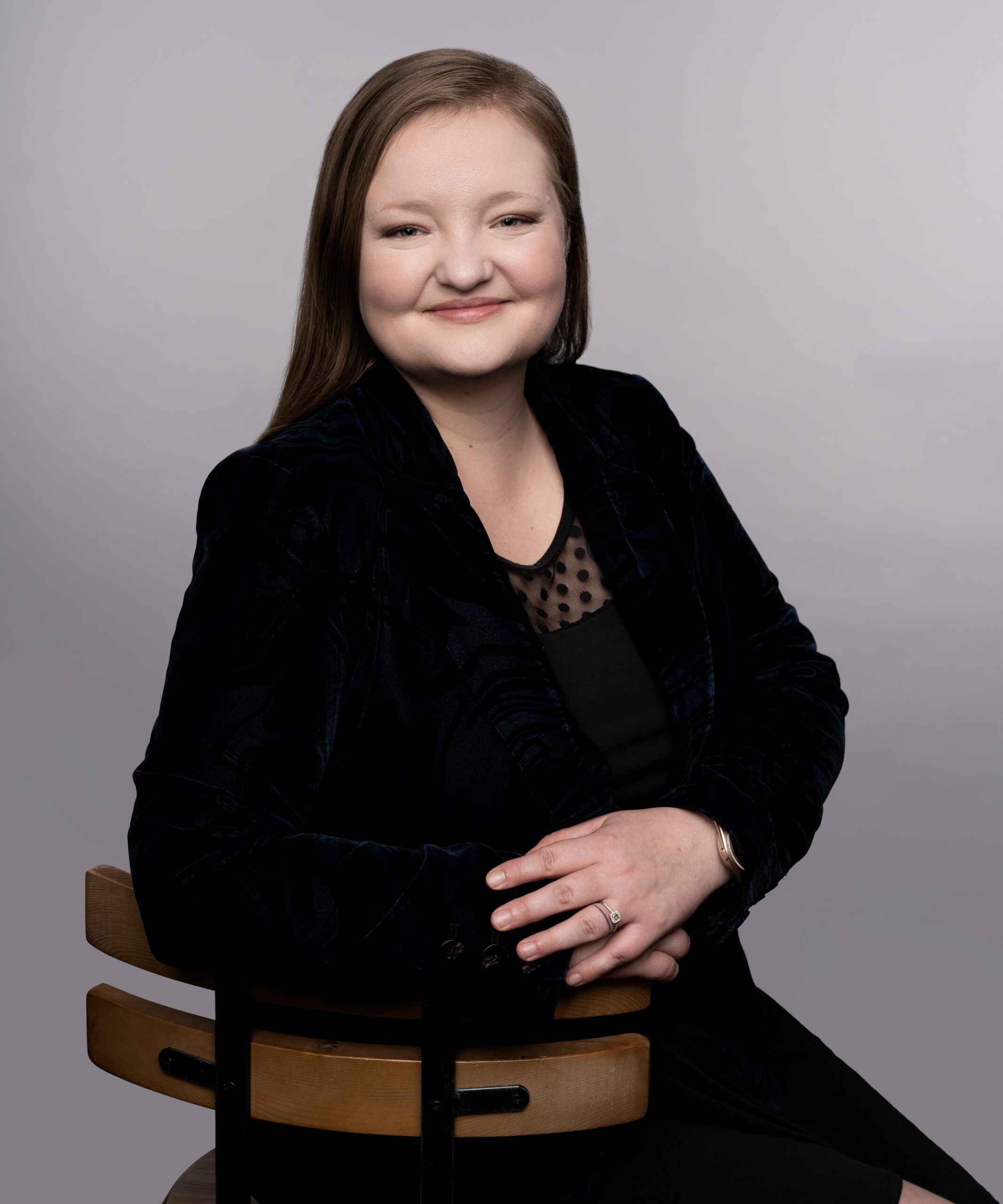Hillary Liljedahl poses confidently for a professional portrait, wearing a dark blazer and seated on a wooden chair, with a neutral background.
