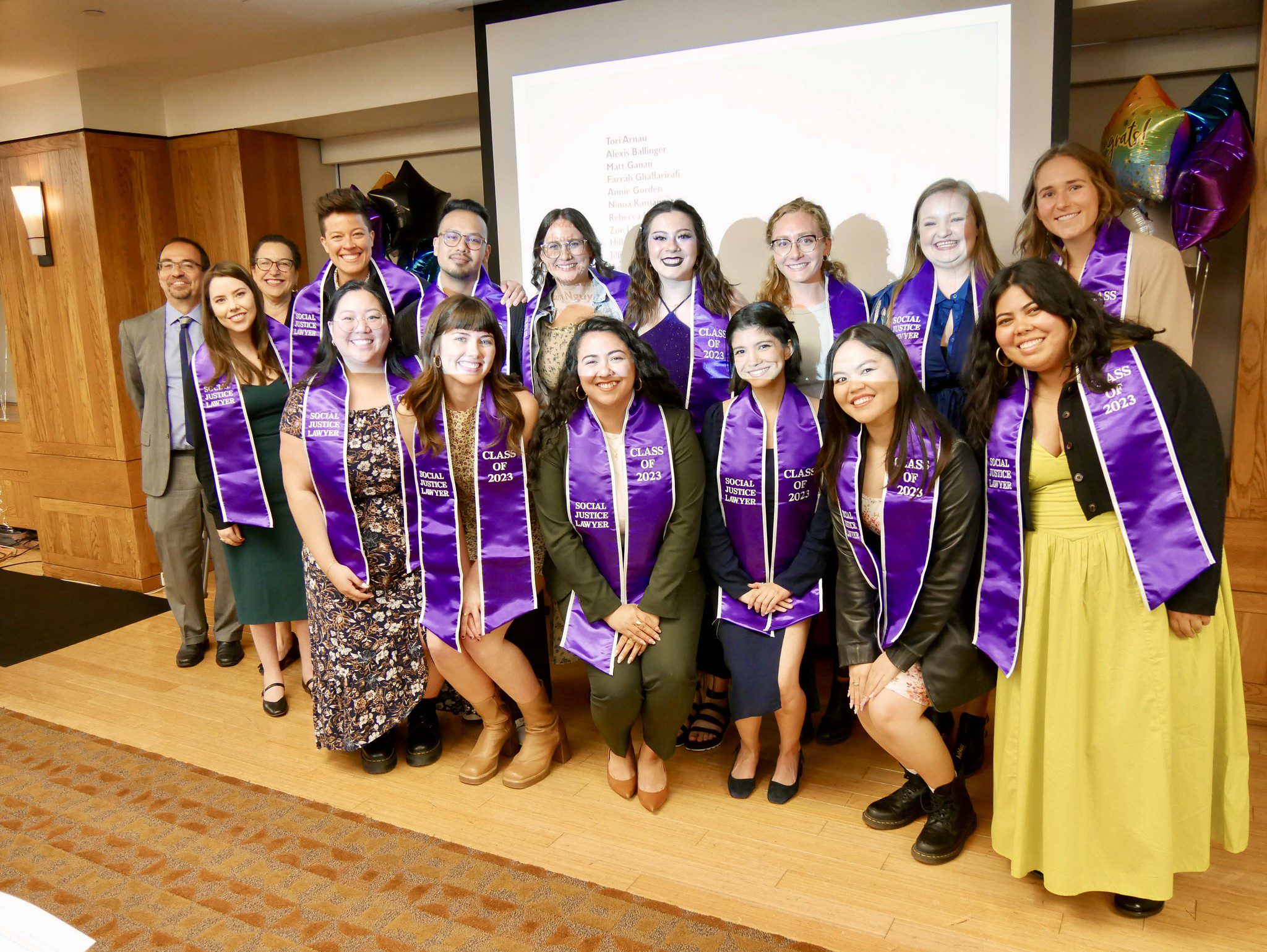 Group of graduates wearing purple stoles for social justice lawyering pose for a photo at a celebration ceremony. A screen and balloons are visible in the background.