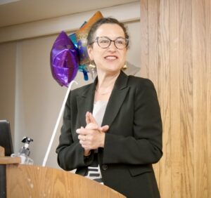 Gail Silverstein speaks at a podium during a ceremony, smiling and clapping her hands, with colorful balloons in the background.