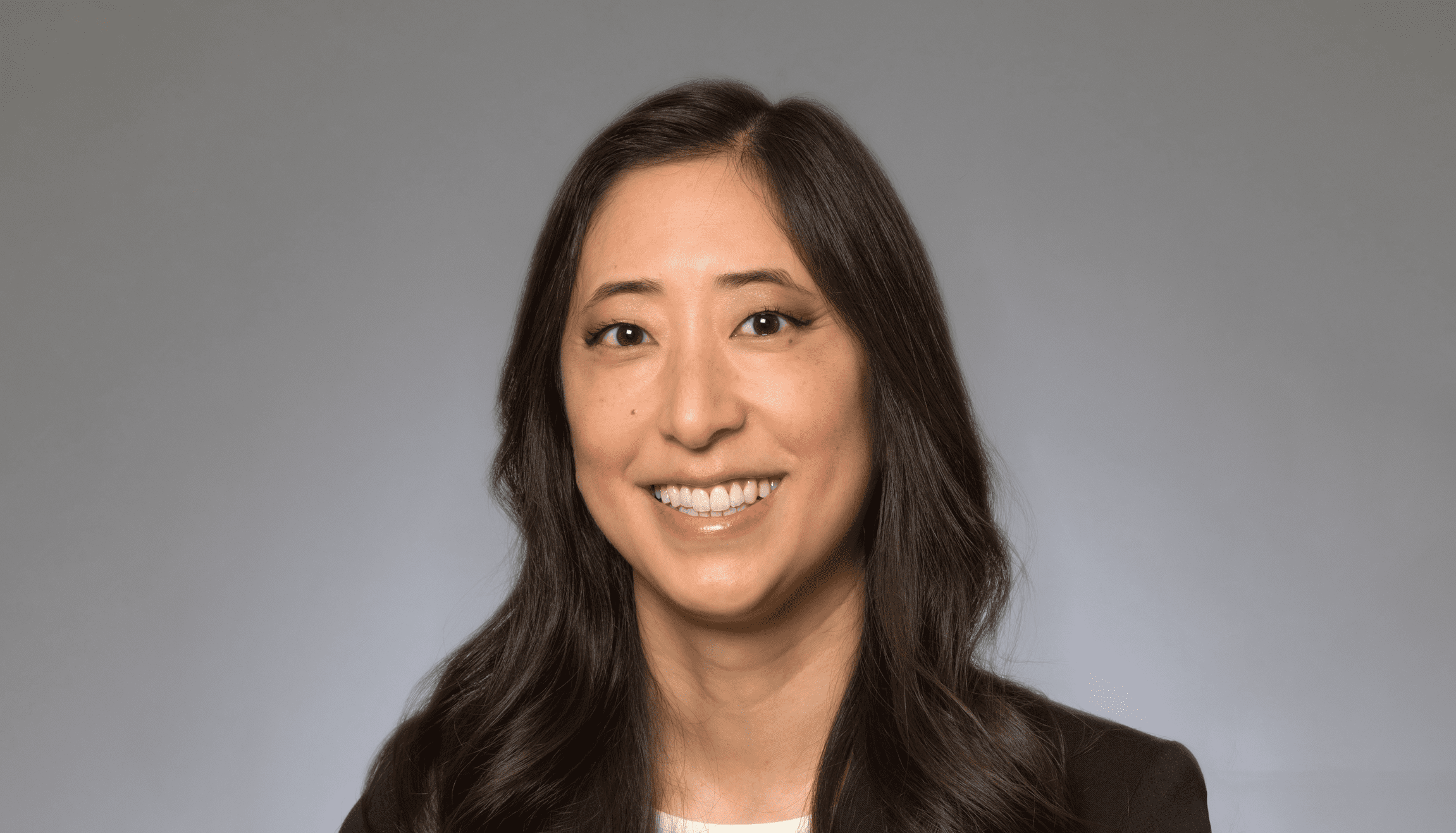 Woman with long brown hair and black suit jacket smiling at the camera. Gray backdrop