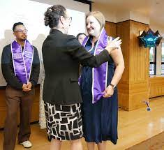 Associate Dean presents a stole to a graduate during a ceremony, with attendees in the background celebrating the occasion.