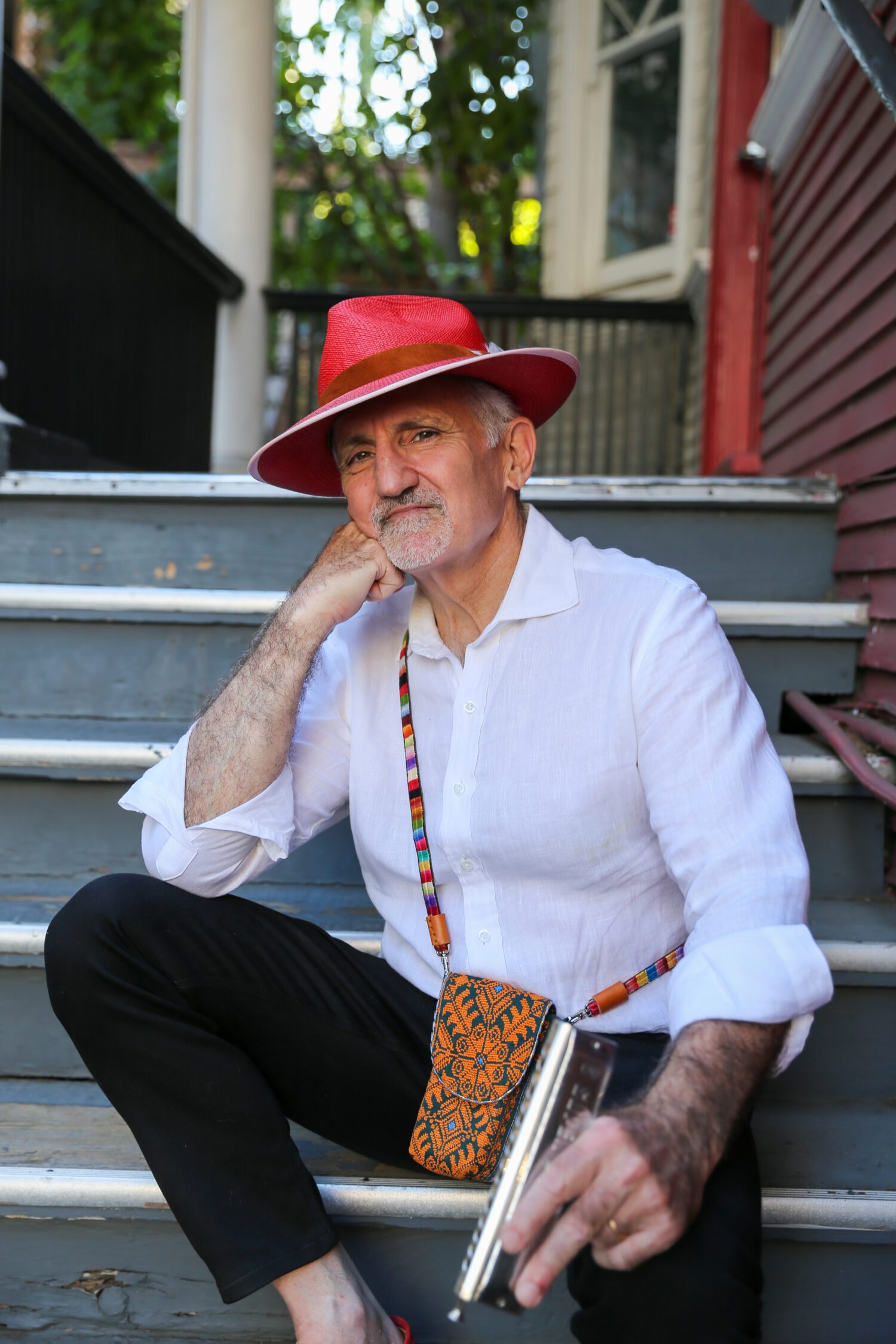 Professor George Bisharat sits on steps wearing a white shirt and a red hat, holding a harmonica and a colorful bag by his side. He looks thoughtfully at the camera.