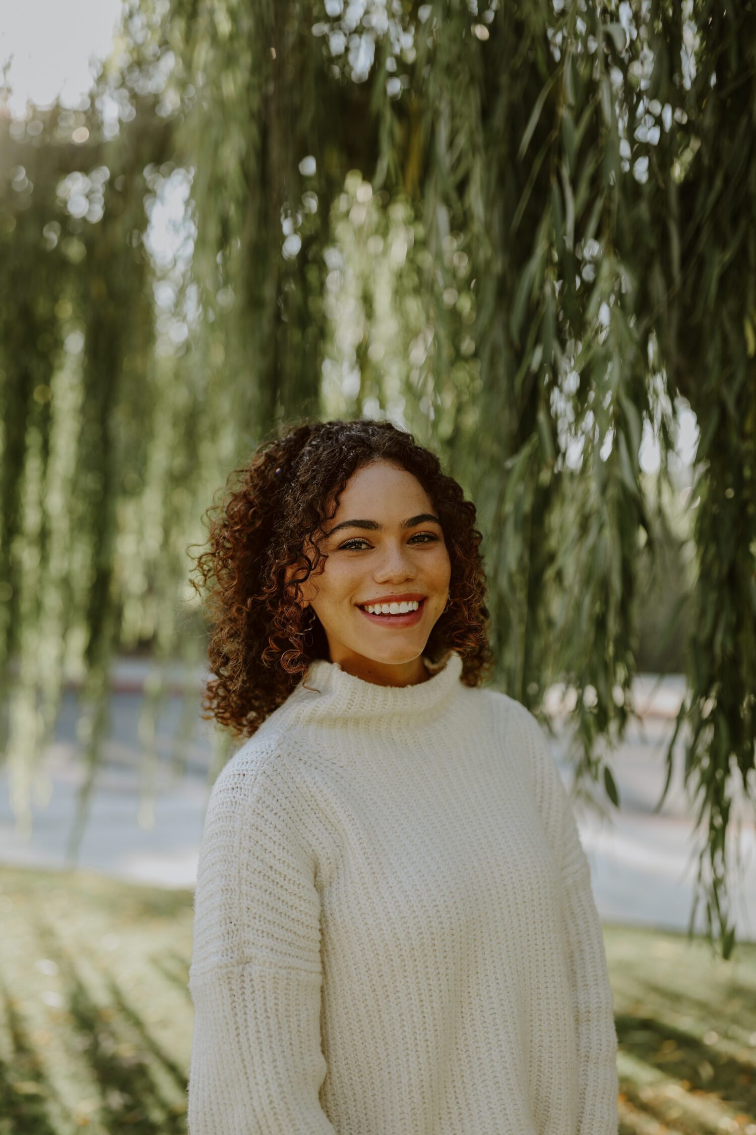 Law student Gabby Olk smiles in a white sweater standing in front of a willow tree