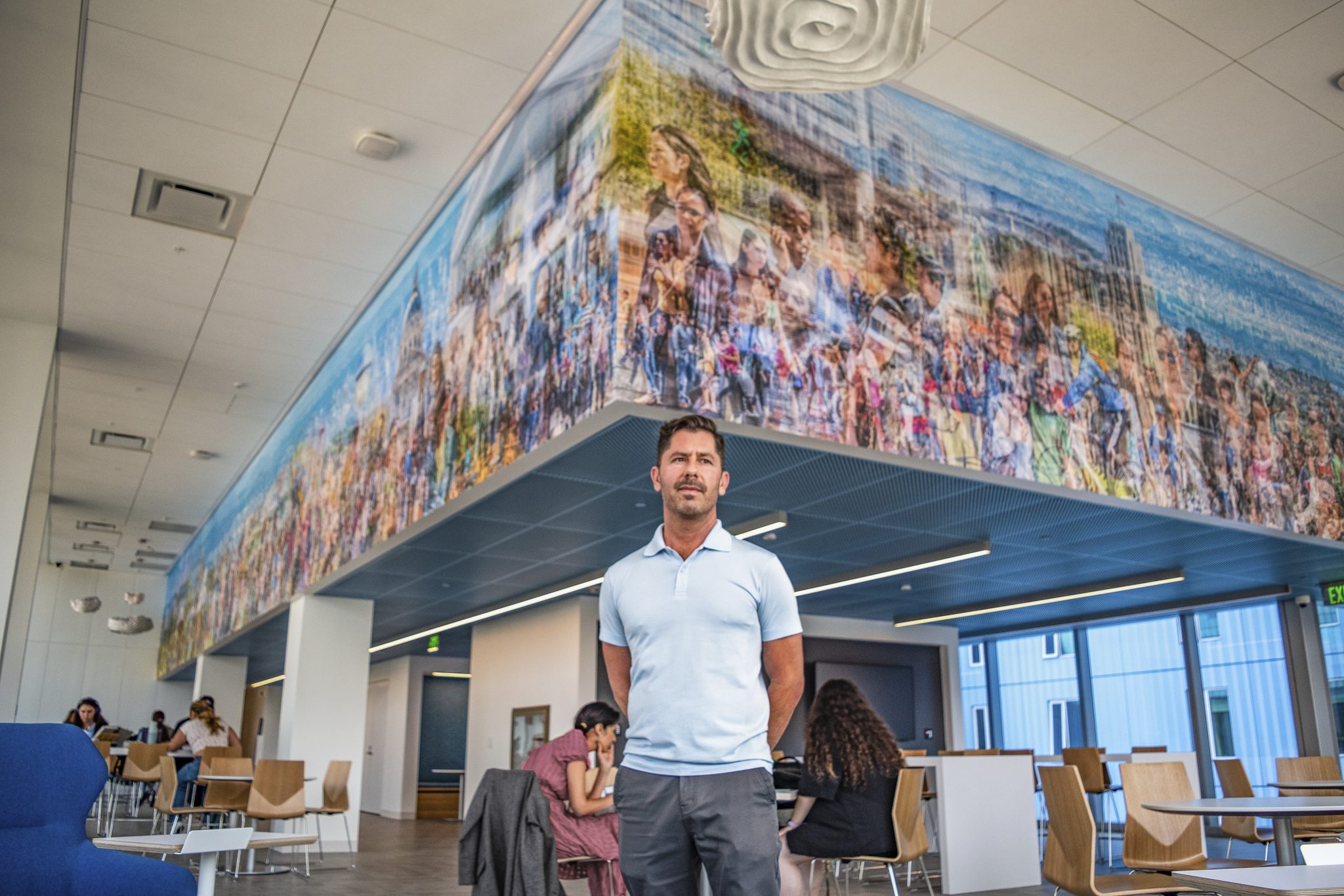Artist and photographer Christopher Dydyk stands in front of his large mural on UC Law SF campus wearing a blue polo shirt and jeans.