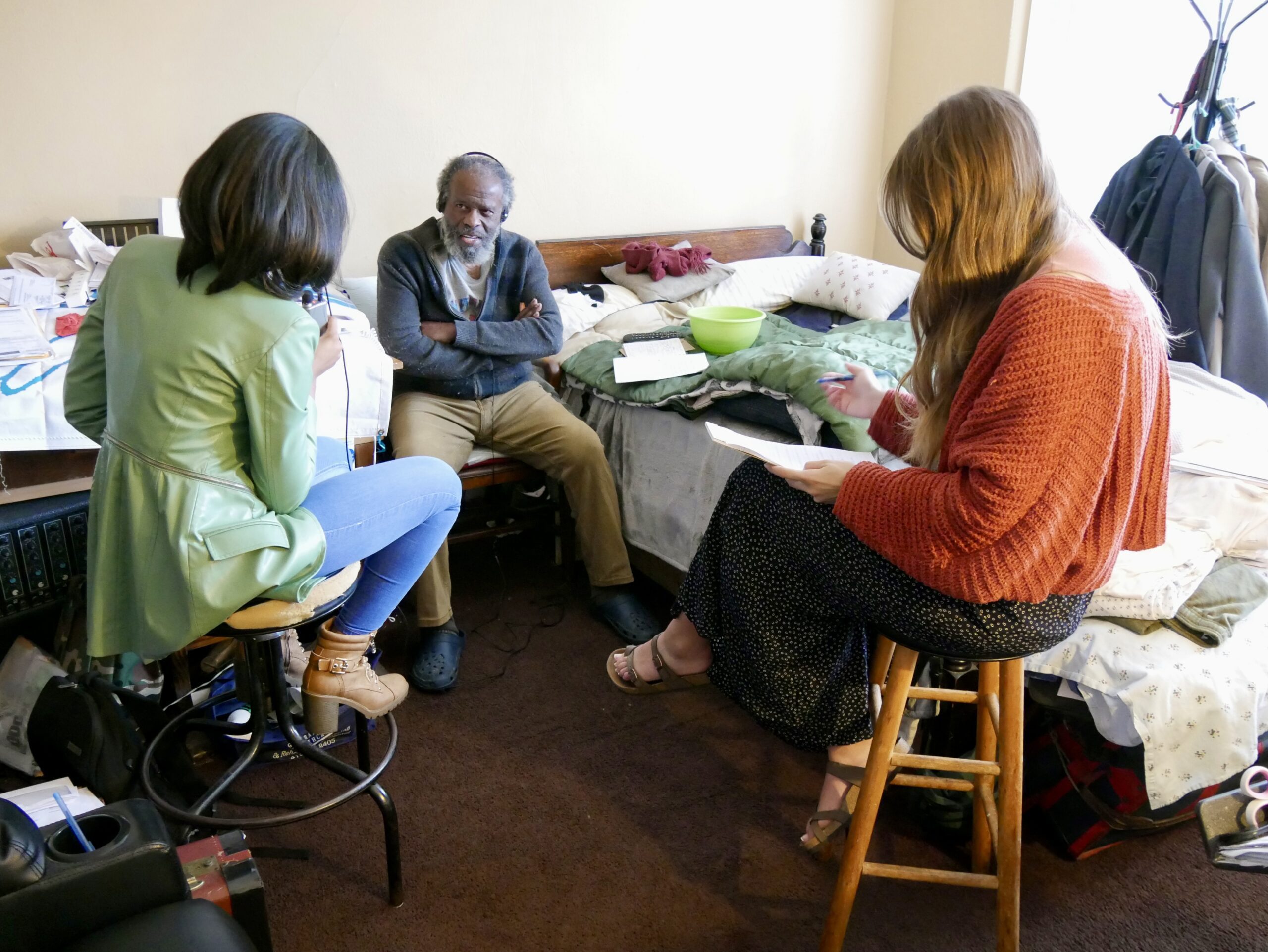 Students Kuwabo Mubyana and Sophia Tarantino are seated and interviewing Patrick Prescod at his bedroom. Prescod is wearing headphones.