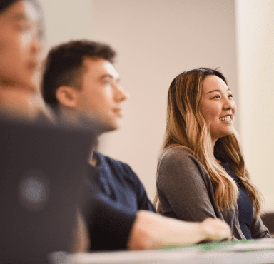Three students in a lecture with the focus on a women student with long hair smiling.