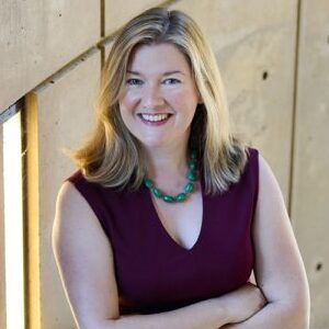 Image of a Bernadette Meyler Carl  smiling with their arms crossed, wearing a burgundy top and green necklace, standing against a concrete wall with light.