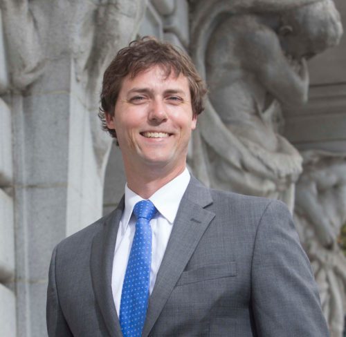 Zachary Price Professor of Law and Eucalyptus Foundation Endowed Chair, UC Law San Francisco in a gray suit and blue tie smiles outdoors in front of a stone building with detailed sculptures, representing a professional setting.