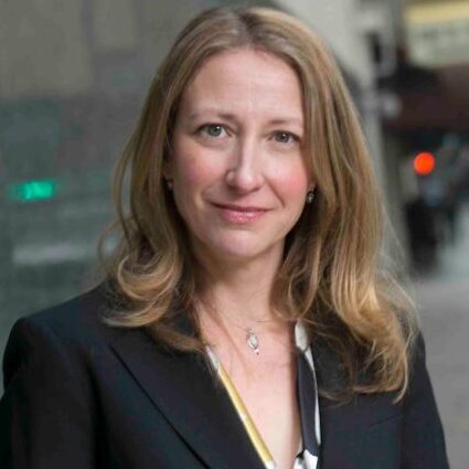 Image of a Jodi Short Mary Kay Kane Professor of Law with long, wavy hair wearing a black suit and a white blouse, smiling and standing outdoors with blurred city buildings in the background.
