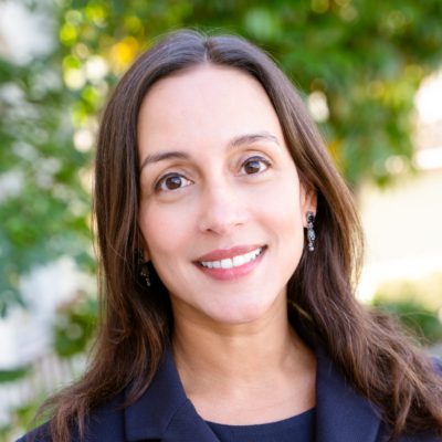 Image of a Mila Sohoni Professor of Law and the John A. Wilson Distinguished Faculty Scholar, Stanford Law School smiling, wearing a dark blazer and earrings, against a blurred outdoor background filled with greenery.