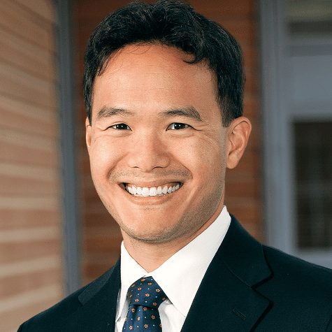 Smiling image of Aaron Tang Professor of Law, UC Davis School of Law in a suit with a white shirt and patterned tie, standing outdoors against a brick background, conveying a professional demeanor.