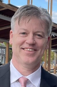 David Makman headshot with salt and pepper hair, a pink tie, and dark suit jacket.