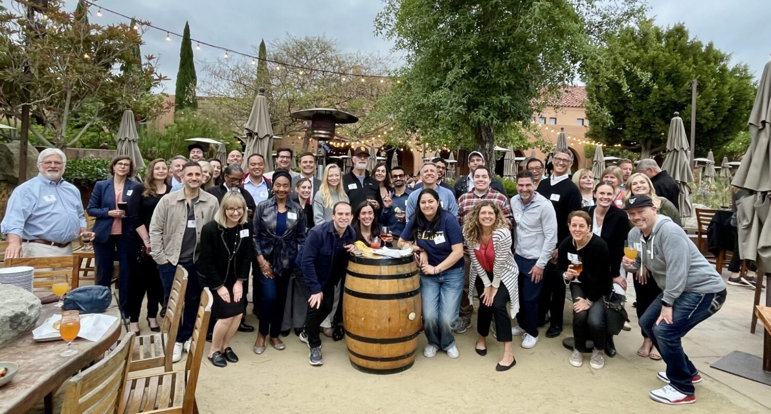 Group photo of attendees at a San Diego Alumni Happy Hour event, gathered outdoors at a venue with drinks and appetizers on tables.