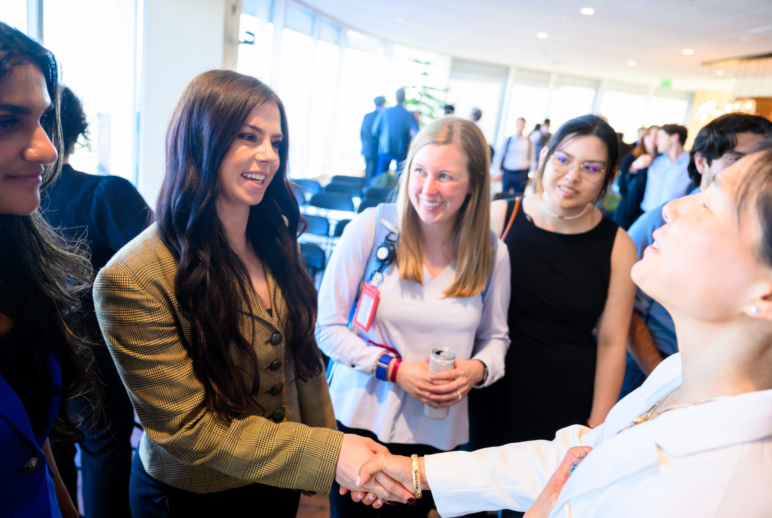 A group of individuals are interacting at an event; one person is shaking hands while others are smiling and engaged in conversation.