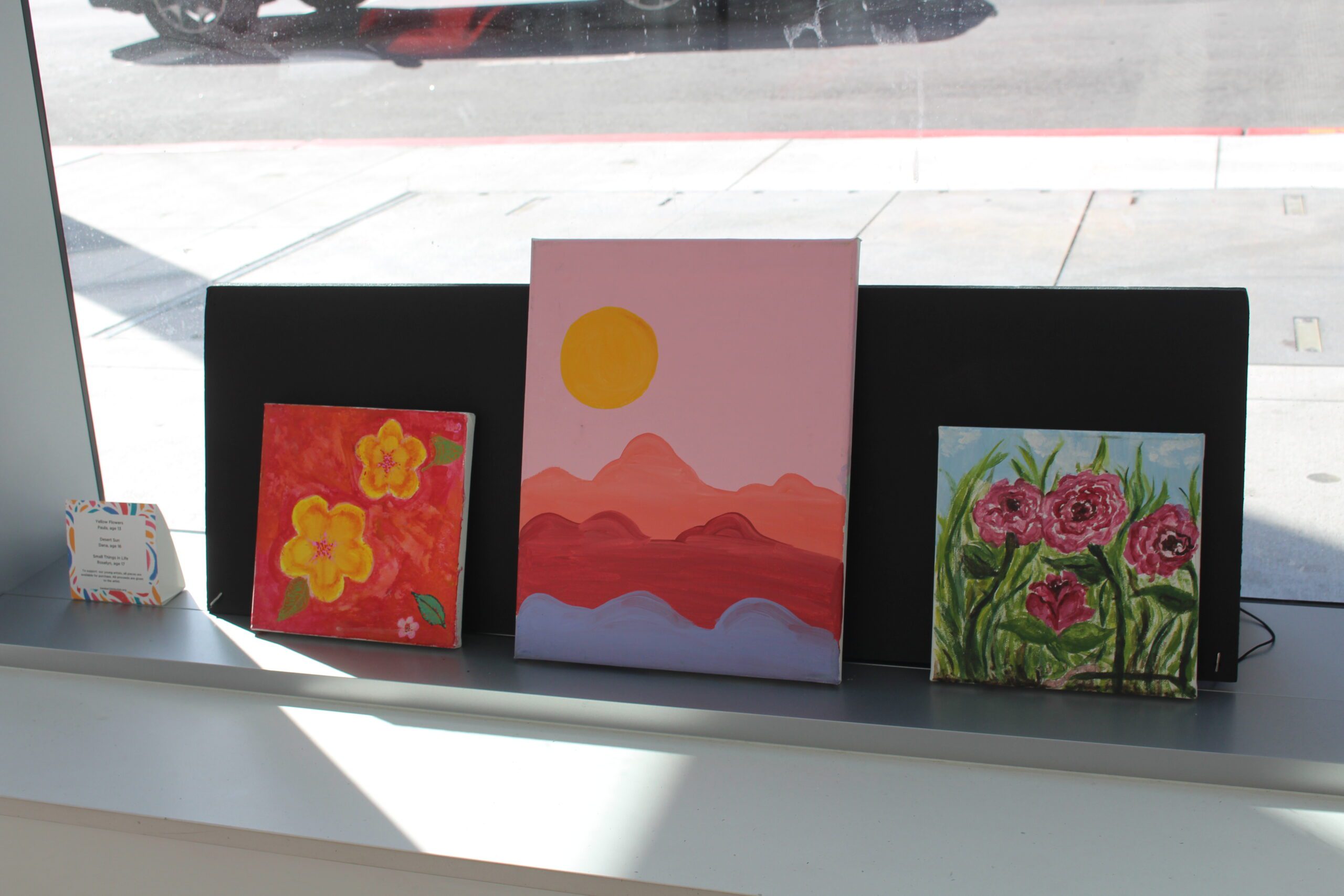 Three paintings on a shelf depicting still lifes and outdoor scenes.