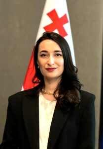 A photo of Mariam Ghvamichava wearing a white shirt and black jacket, standing in front of a red and white flag.