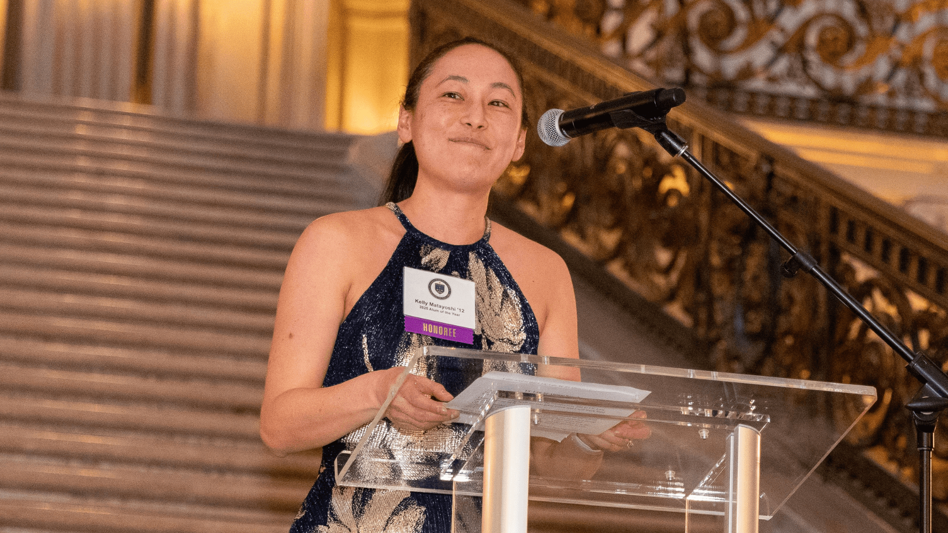 2025 Alum of the Year Kelly Mataoyoshi stands behind a lecturn at San Francisco City Hall during an award reception.