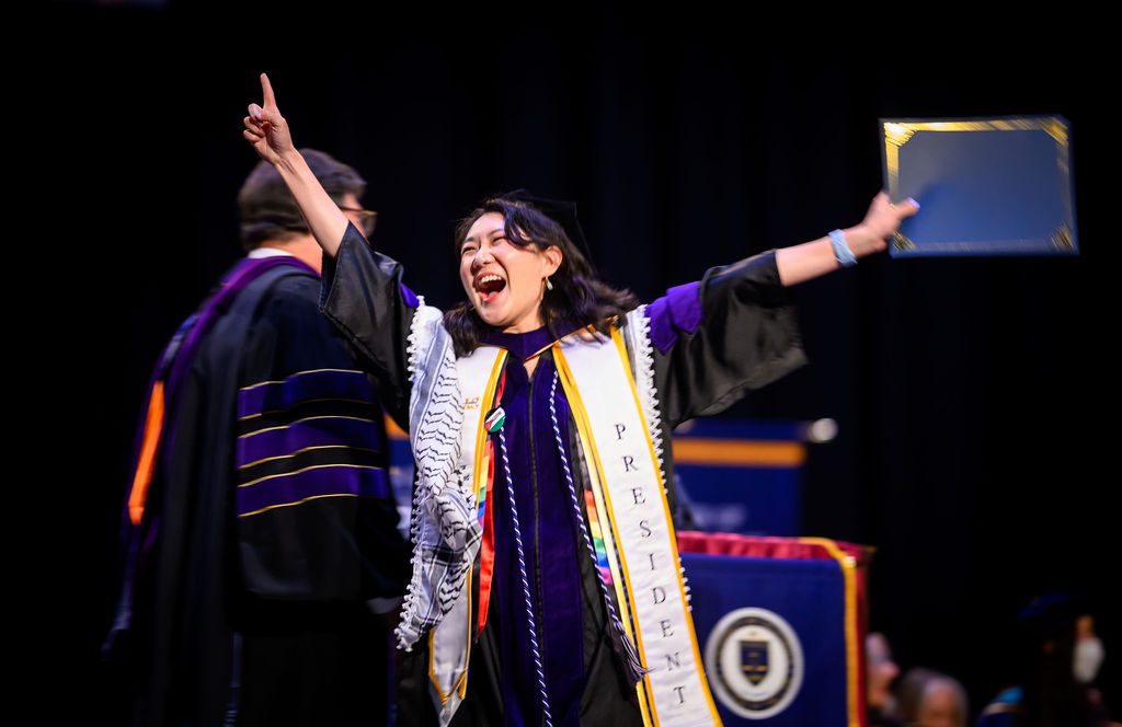 A graduate in a cap and gown and arms in the air celebrates receiving her degree on stage.
