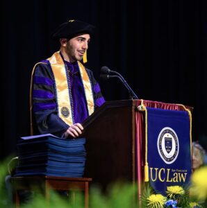 Paolo Maggisano LLM ’25 speaks behind a lecturn wearing a black robe and gold stole