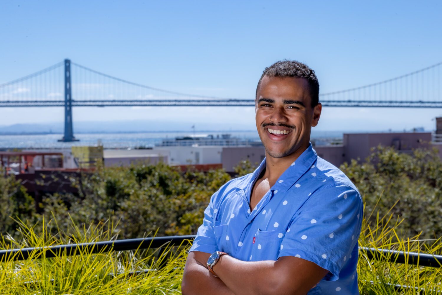 photo of Mayell Mergaert posing outside in a blue short-sleeved shirt with arms crossed with the bay and a bridge behind him