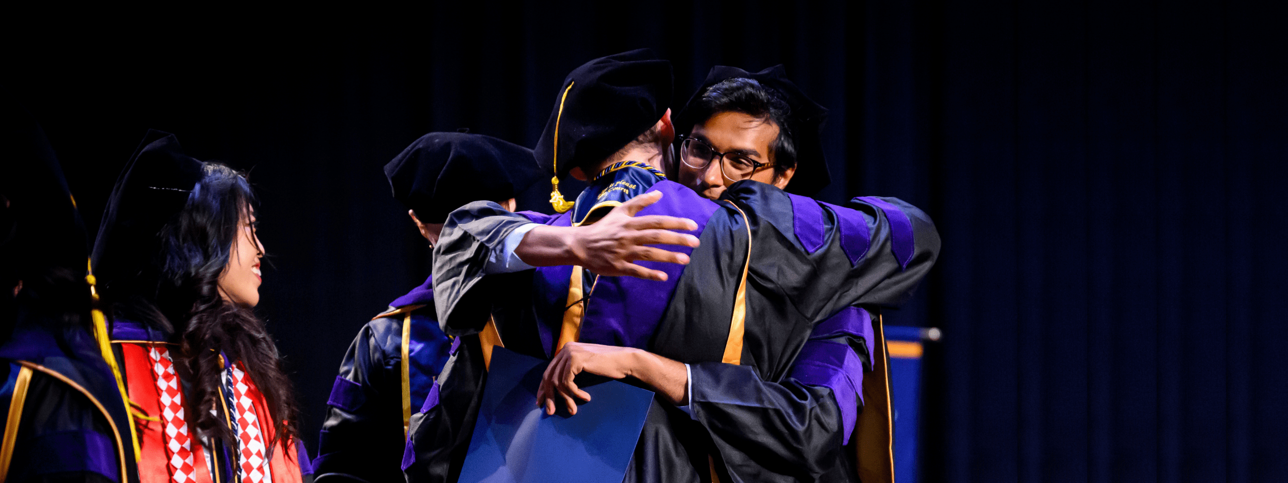 Two members of the Class of 2025 dressed in graduation regalia hugging on stage during commencement