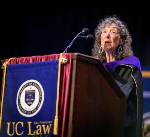 Professor Karen Musalo wearing a black gown while speaking behind a lecturn with a UC Law SF banner