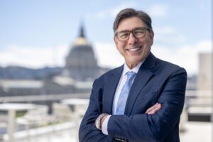 A portrait of David Faigman. He is smiling and looking at the camerea. His arms are crossed and he wears a blue suit. City Hall can be seen in the background.