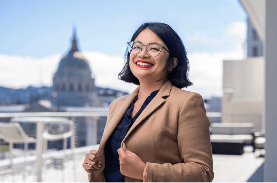 A headshot of Ashley Judilla wearing a blazer and glasses. City Hall can be seen in the background