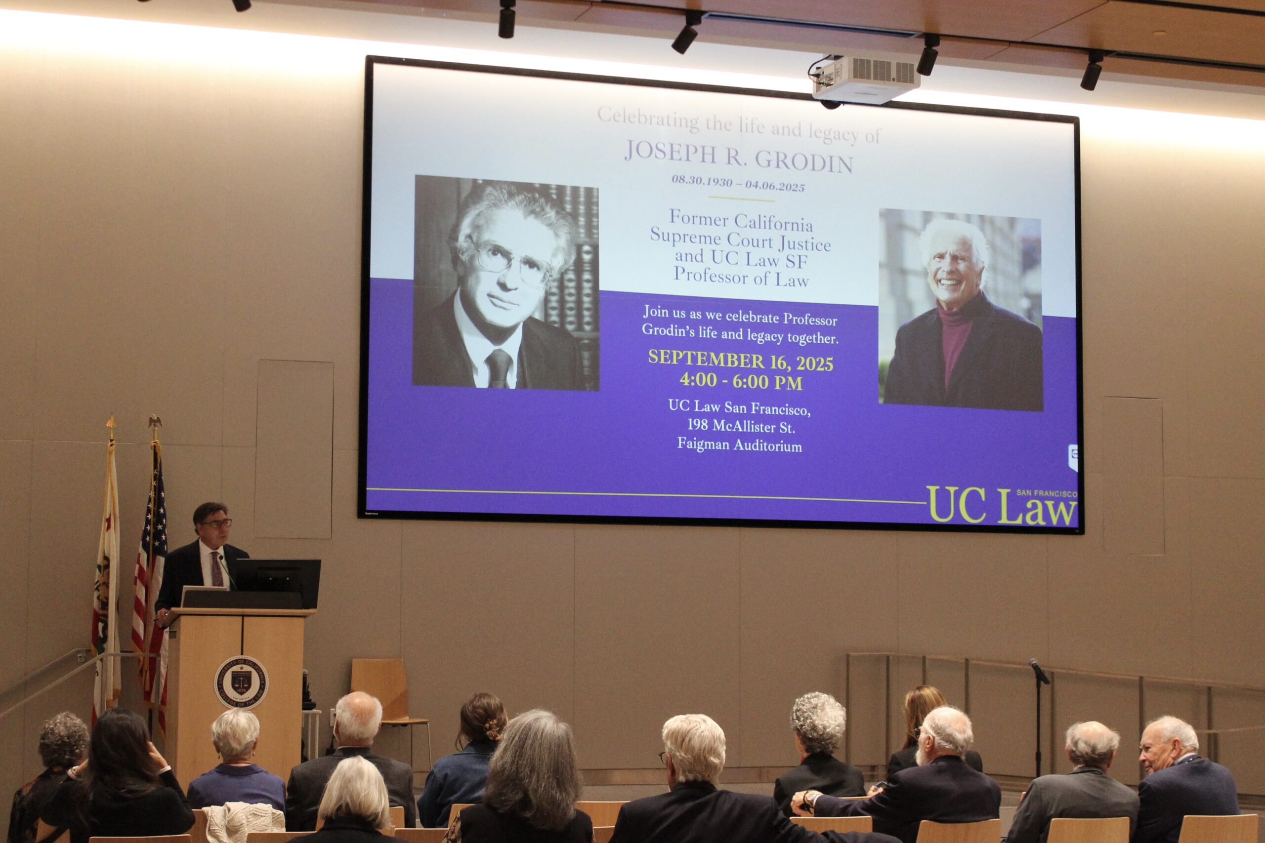 David Faigman speaks at a podium in front of an audience beside a screen displaying photos of Joseph Grodin.