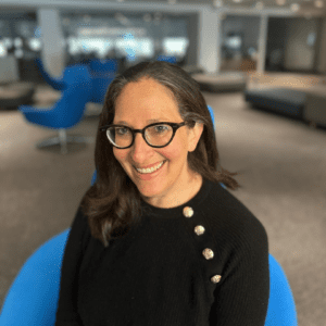 Nicole Ozer smiling and looking at the camera. She sits on a blue chair and wears a black sweater with gold buttons. She also wears glasses and her long hair is down.