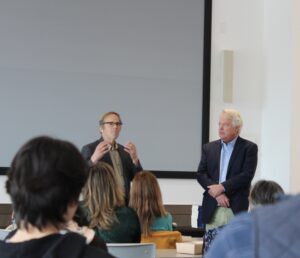 Randy Shaw and David Seward speak at the front of a room before an audience.