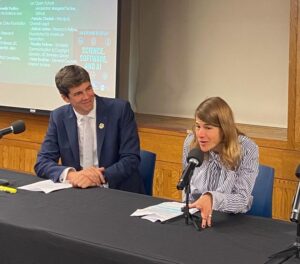 French Consul General Florian Cardinaux, seated wearing a suit, watches Tal Niv speak from a table with microphones.