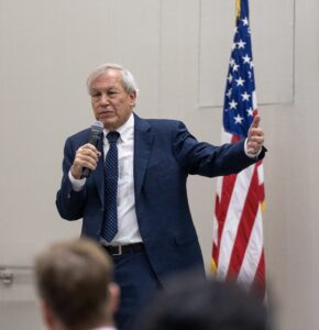 Erwin Chemerinsky speaks wearing a blue suit with an american flag in the background.