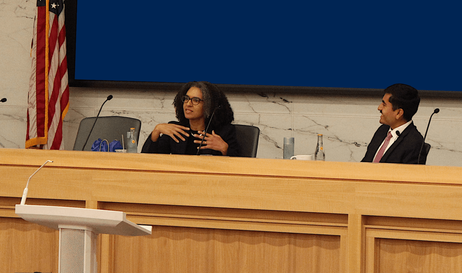 Leondra Kruger speaks behind a bench in a mock courtroom sitting next to a law student with an American flag in the background.