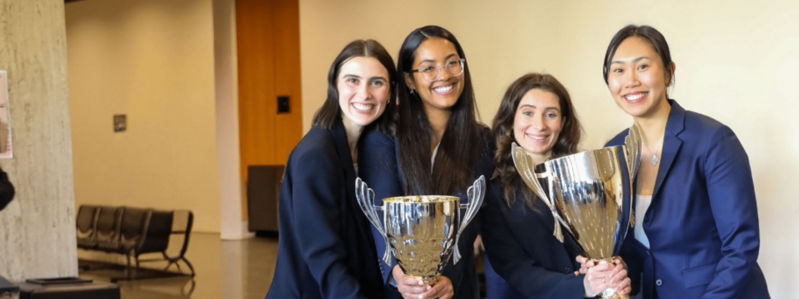 Four students in business attire stand next to each other smiling. They are holding two large gold trophies.