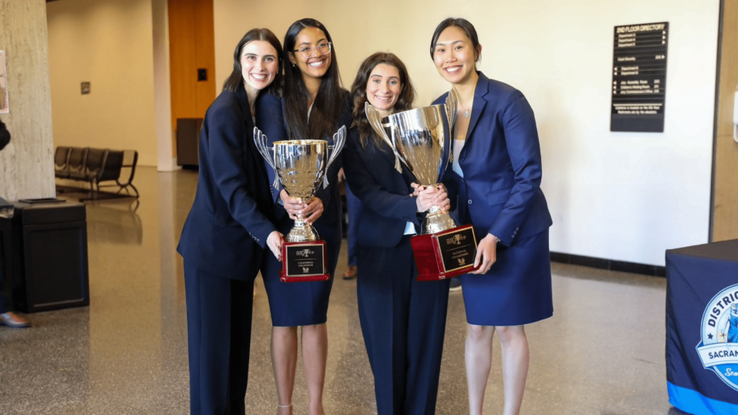Four students in business attire stand next to each other smiling. They are holding two large gold trophies.
