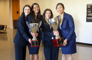 Four students in business attire stand next to each other smiling. They are holding two large gold trophies.