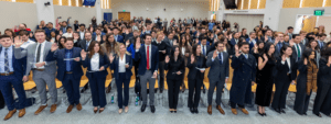 A large group of graduates raise their right hands in the air during a swearing-in ceremony in a packed auditorium.