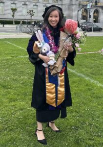 Stella Low wears a black cap and gown for graduation while standing on a green lawn in front of San Francisco City Hall.