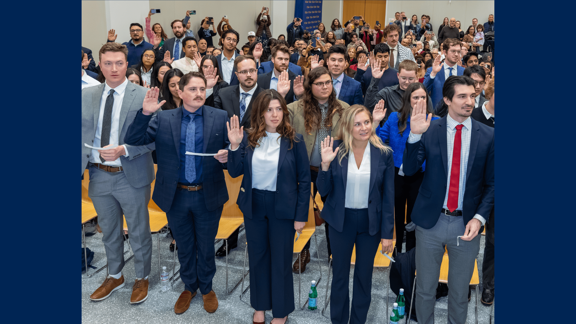 A large group of graduates raise their right hands in the air during a swearing-in ceremony in a packed auditorium.