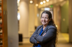 Dorit Reiss smiling in the UC Law SF library. Her arms are crossed, and she's wearing a gray blazer over a blue shirt.