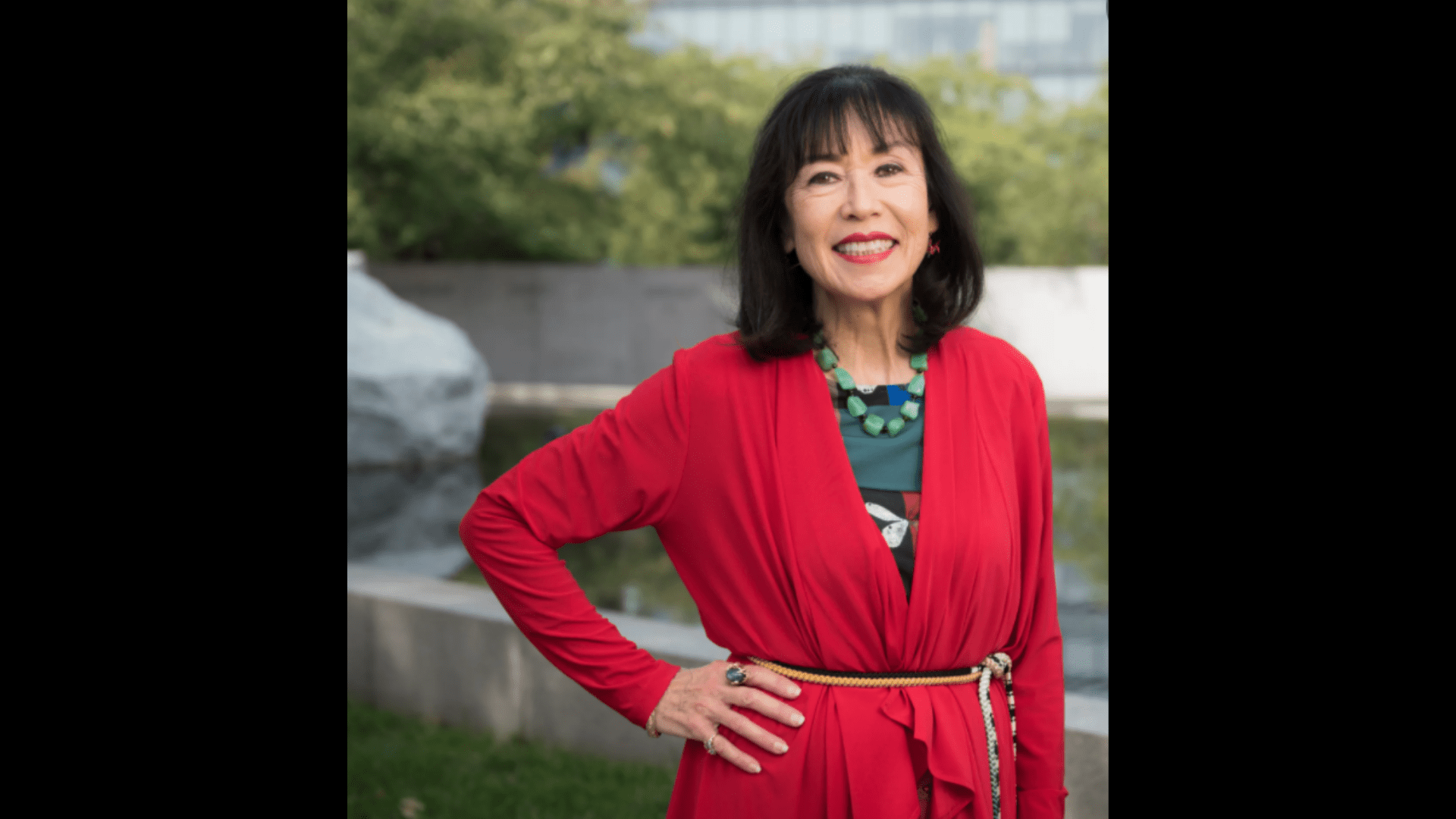 Karen Korematsu, with dark hair, poses outdoors in a red dress, hand on her hip.