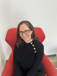 Nicole Ozer looks at the camera wearing glasses and a dark shirt, sitting on a red chair.