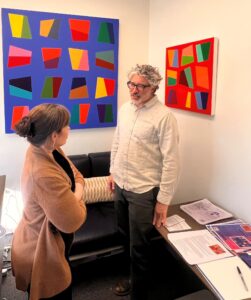 Pablo Manga speaks with an individual in an office where two pieces of his art adorn the wall.