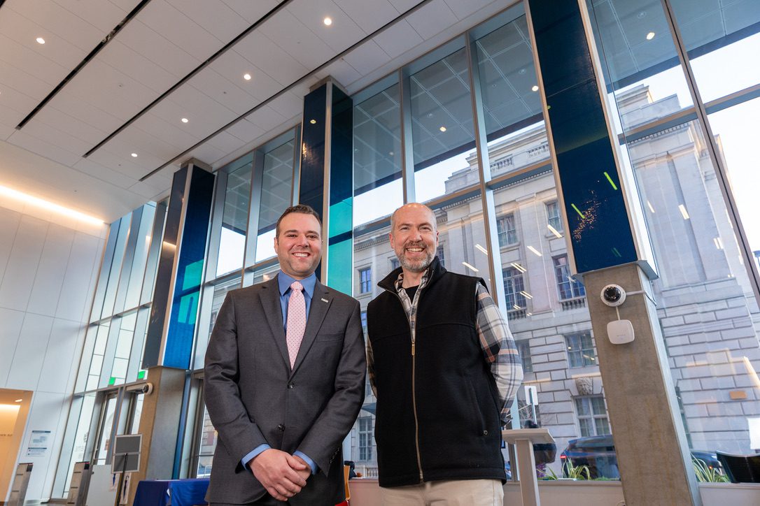 Dan Goods and Christopher Gabriel stand in the lobby of the 198 McAllister building, a digital art installation behind them.
