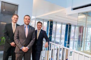 Engineers Dylan Lukes, Christopher Gabriel, and Kaden Neary pose in front of digital artwork displayed on three pillars in a building lobby.