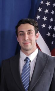 Headshot of law student Ediz Kalkan wearing a suit and tie, American flag behind him.