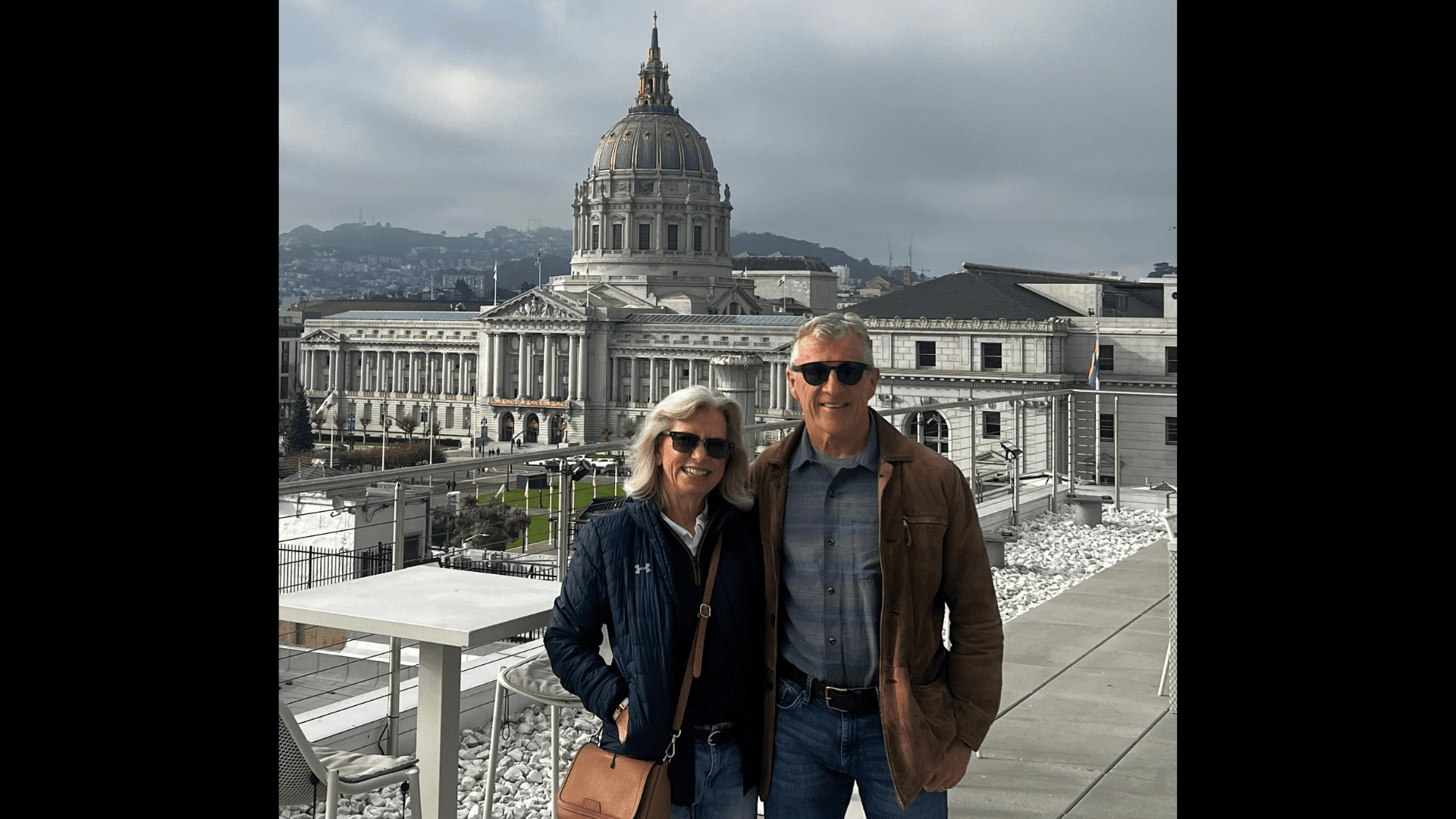Jennifer and Bob Hagle, wearing sunglasses and warm clothes, pose in front of the Sky Deck of 333 Golden Gate Ave. with the San Francisco City Hall dome and building behind them.