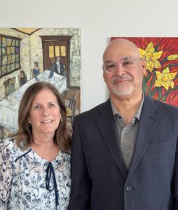 Jo Ann and Michael Montoya pose for a photo, a wall with paintings behind them.